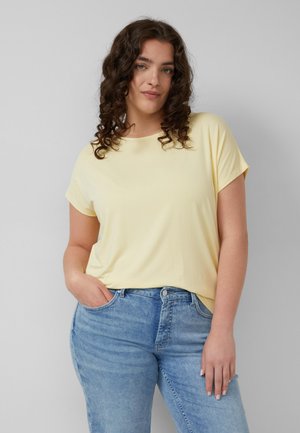 Frau mit lockigem dunklem Haar, die ein blassgelbes kurzärmeliges Oberteil und hellblaue Jeans trägt, posiert mit einer Hand in der Tasche vor grauem Hintergrund.