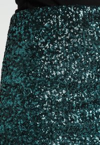 Tessuto in lurex turchese, densamente coperto da cirri di brillanti paillettes circolari, crea una superficie testurizzata. Lo sfondo è scuro, esaltando la lucentezza delle paillettes.