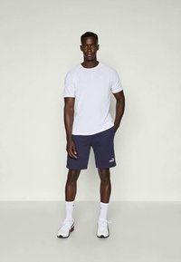 Fehér rövid ujjú póló és tengerészkék rövidnadrág fehér logóval. A modell fehér sportcipőt visel fekete részletekkel. Egyszerű, sportos dizájn.