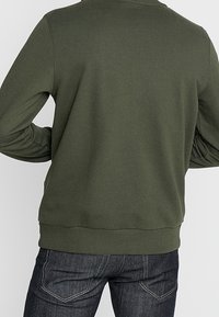 Felpa verde oliva con scollo rotondo, polsini e fondo a costine. Abbinata a jeans in denim scuri con cuciture a contrasto e tasche posteriori.