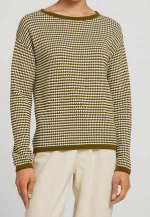 Personne portant un pull à manches longues à carreaux vert olive et blanc, associé à un pantalon beige clair, debout devant un fond uni.