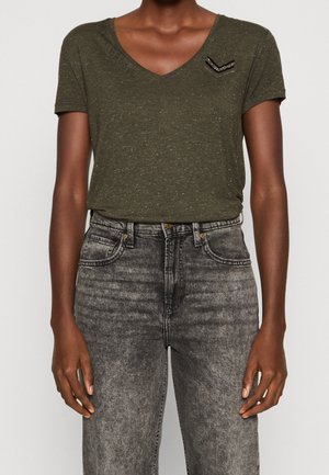 Personne portant un T-shirt vert olive à col en V avec un petit écusson chevron noir et argenté, et un jean noir délavé taille haute.