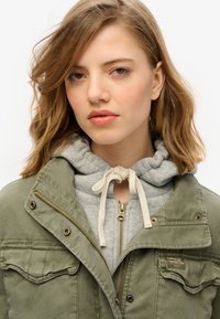 Olivgrön jacka med dragkedjefickor, tryckknappar och synlig sömnad, lager på lager över en grå huvtröja med snörning och dragkedja.