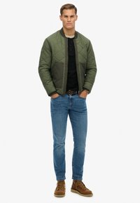 Groene gewatteerde bomberjack met effen mouwen, zwart henley shirt, blauwe spijkerbroek en bruine veterschoenen. Gestyled met een horloge.