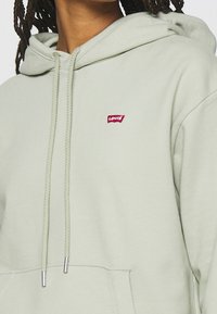Jasnozielona bluza z kapturem wykonana z miękkiego materiału, z kieszenią z przodu oraz małym czerwonym logo Levi's na piersi.
