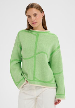 Ženska, ki nosi svetlo zeleni pulover z vzorcem in bele hlače, ki pozira z eno roko v žepu na enobarvnem ozadju.