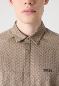 Trumpas rankovės, rudos spalvos raštuotas marškinėlis su mygtuku ir subtiliu geometriniu dizainu. Krūtinėje yra juodas "BOSS" logotipas.