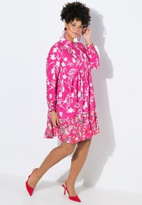 Rosa Blumenkleid mit langen Ärmeln, einem runden Kragen und einem gestuften Rock. Verfügt über weiße Blumenmuster und knallrote High Heels.