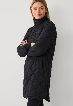 Vrouw met licht haar die een lange zwarte doorgestikte jas met zijzakken draagt en met gekruiste armen staat tegen een effen achtergrond.