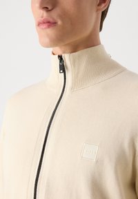 Sweatshirt beige zippé avec col montant, tissu texturé et fermeture éclair noire. Présente un logo en relief subtil sur la poitrine.