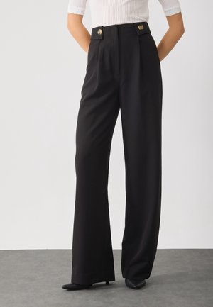 Frau trägt schwarze, weit geschnittene Hose mit hoher Taille und Knopfdetails, kombiniert mit einem weißen, gerippten Kurzarmoberteil und schwarzen, spitzen Schuhen.
