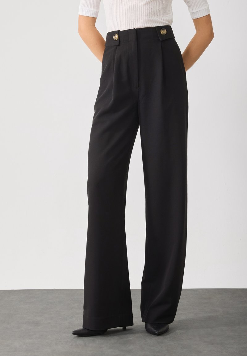 Femme portant un pantalon large noir taille haute avec des détails de boutons, associé à un haut blanc côtelé à manches courtes et des chaussures noires à bouts pointus.
