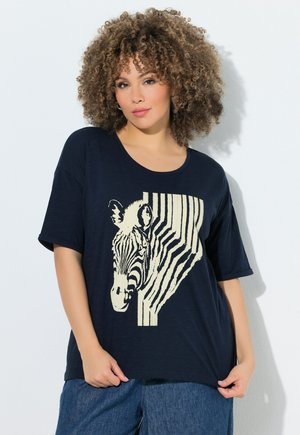 Mujer con cabello rizado que lleva una camiseta azul marino con un diseño de media cara de cebra y rayas, combinada con vaqueros azules.