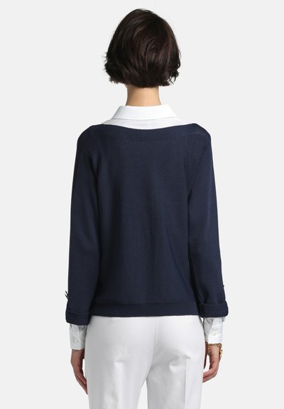 Pulover bleumarin tricotat, cu croială lejeră, guler larg și manșete răsucite, purtat peste o cămașă albă cu guler, cu material texturat.