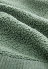 Groene fleece stof, textuur met een zachte, vlezige oppervlakte. Het materiaal vertoont lichte plooien en heeft een geribbelde randafwerking.