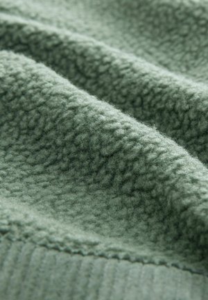 Green fleece fabric, textured with a soft, fuzzy surface. The material shows gentle folds and a ribbed edge detailing.
