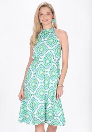 Tânără zâmbitoare, purtând o rochie albă fără mâneci, de lungime până la genunchi, cu modele geometrice verzi și albastre și cercei turcoaz.