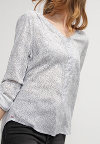 Jasnoniebieska bluzka z dekoltem w serek, długimi, podwiniętymi rękawami, wzorem w grochy oraz guzikami z przodu. Miękka faktura materiału.