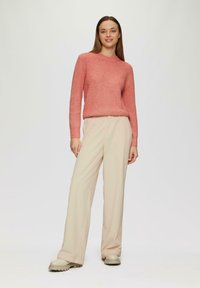 Roze ribbeltrui met een ronde hals, gecombineerd met beige wijde broek en crèmekleurige chunky schoenen. Model staat tegen een effen achtergrond.