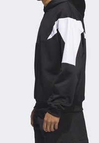 Homme portant un sweat à capuche noir avec de grands panneaux géométriques blancs sur le haut des manches, debout de profil sur un fond uni.