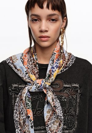 Joven con cabello corto y castaño que lleva una colorida bufanda floral atada sobre una blusa oscura con bordados y un largo pendiente dorado colgante.