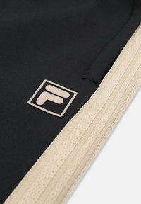 Primo piano di un tessuto nero con bordatura lavorata a maglia beige e una toppa con logo ricamato beige che ricorda la lettera "F" all'interno di un quadrato.