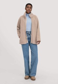 Giacca in pile beige con colletto alto, abbinata a un dolcevita azzurro chiaro e jeans in denim a vita alta. Presenta una vestibilità relax e un design minimale.