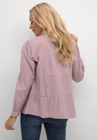 Blusa rosa chiaro con design a peplo, caratterizzata da maniche lunghe e dettagli arricciati lungo lo yoke e l'orlo. Tessuto dalla texture liscia.