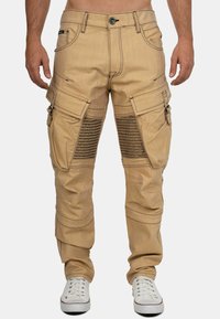 Pantalons cargo beiges avec de grandes poches latérales, accents de design côtelés et couture contrastante, fabriqués en tissu robuste. Portés avec des baskets blanches.