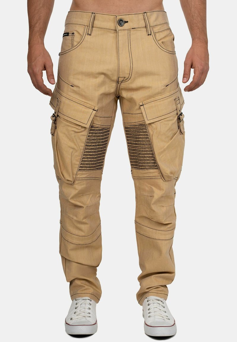 Pantalons cargo beiges avec de grandes poches latérales, accents de design côtelés et couture contrastante, fabriqués en tissu robuste. Portés avec des baskets blanches.