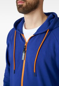 Blue zip-up hoodie featuring orange drawstrings and zipper. Fabric texture is smooth with a relaxed fit. Logo tag is visible near the zipper.