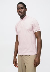 Šviesiai rožinė polo marškinėliai, siauro kirpimo, su trimis sagutėmis ir mažu logotipo lipduku. Turi kontrastuojančias baltas apykaklės ir rankovių detales.