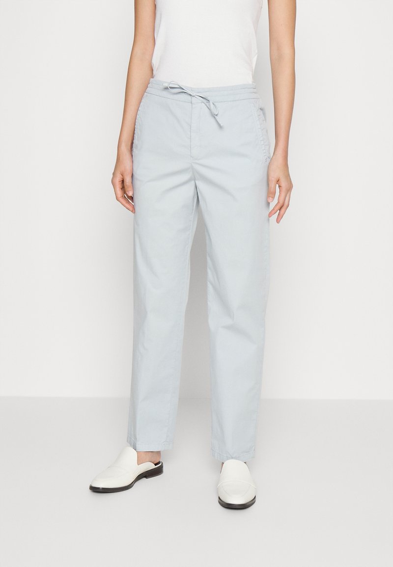 Femme portant un pantalon droit gris clair avec une taille à cordon et des chaussures blanches sans lacets, debout devant un fond uni.