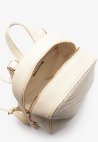 Krém színű műbőr hátizsák szőtt anyagú elülső résszel, dupla cipzáras rekeszekkel és arany színű kiegészítőkkel. Állítható pántok tartoznak hozzá.