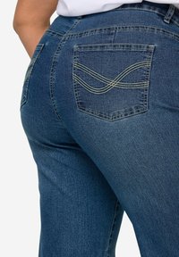 Vaqueros de mezclilla en azul oscuro con una textura suave. Cuentan con bolsillos traseros con detalle de costura amarilla y una silueta ajustada.