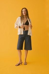 Jeune femme portant un haut de bikini noir, un cardigan blanc en maille ouverte, un short bermuda à fines rayures et des sandales à talons noires sur fond jaune.