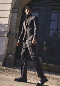Traje gris oscuro con un patrón texturizado, chaqueta ajustada y pantalones estrechos. Combinado con zapatos negros y un gorro de punto negro. Entorno urbano.