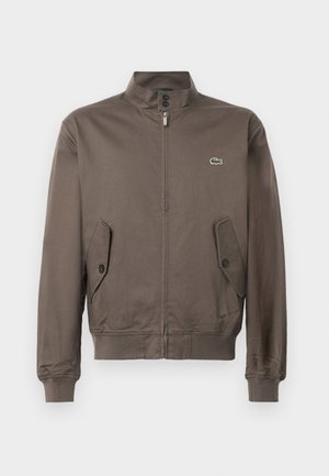 Bruine ritssluitingjack met staande kraag, knoopflapzakken, ribgebreide manchetten en onderrand, en een klein groen krokodillenlogo op de borst.