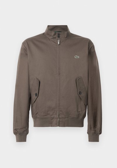 Veste zippée marron avec col montant, poches à rabat boutonnées, bords côtelés aux poignets et à l'ourlet, et petit logo de crocodile vert sur la poitrine.