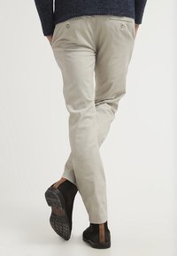 Persona che indossa pantaloni beige e stivali Chelsea marrone scuro, che cammina con le mani in tasca su uno sfondo neutro.
