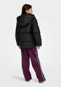 Crna prozračna jakna s kapuljačom, debeli jastučićasti dizajn s teksturiranim završetkom, uparena s ljubičastim hlačama koje imaju bočne pruge.