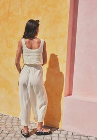 Cream crochet crop top paired with loose, light-colored culottes and dark sandals, set against a vibrant yellow and pink wall.