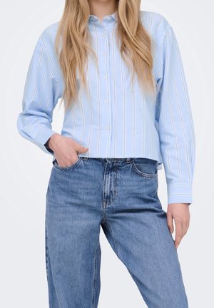 Frau trägt ein hellblau gestreiftes Hemd mit Knöpfen und hoch taillierte blaue Jeans, mit einer Hand in der Tasche und langen blonden Haaren.