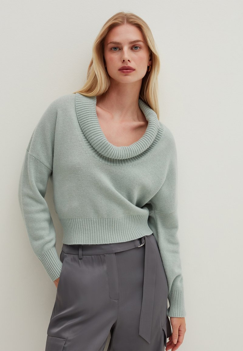 Mintgrüner Pullover mit Schalkragen aus weichem, glattem Material, mit geripptem Saum und lockerer Passform, kombiniert mit grauen Satin-Hosen und einem gebundenen Detail an der Taille.