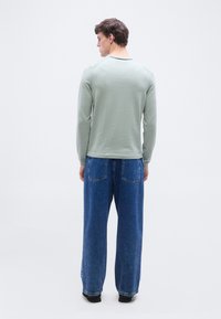 Άνδρας όρθιος, γυρισμένος πλάτη, φοράει ανοιχτό πράσινο πουλόβερ, μπλε τζιν παντελόνια φαρδιάς γραμμής και μαύρα παπούτσια, με φόντο λευκό απλό.