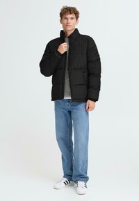 Sort pufferjakke med lynlås foran, stående krave og elastiske manchetter, parret med en grå sweater, blå jeans og hvide sneakers.