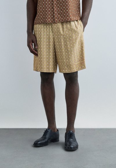 Homme portant un short beige à motifs et une chemise marron à motifs associée à des chaussures de ville noires, debout sur un sol gris devant un fond clair.