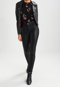Woman wearing black leather jacket, black floral top, black leather pants, and black ankle boots, standing against a plain background.