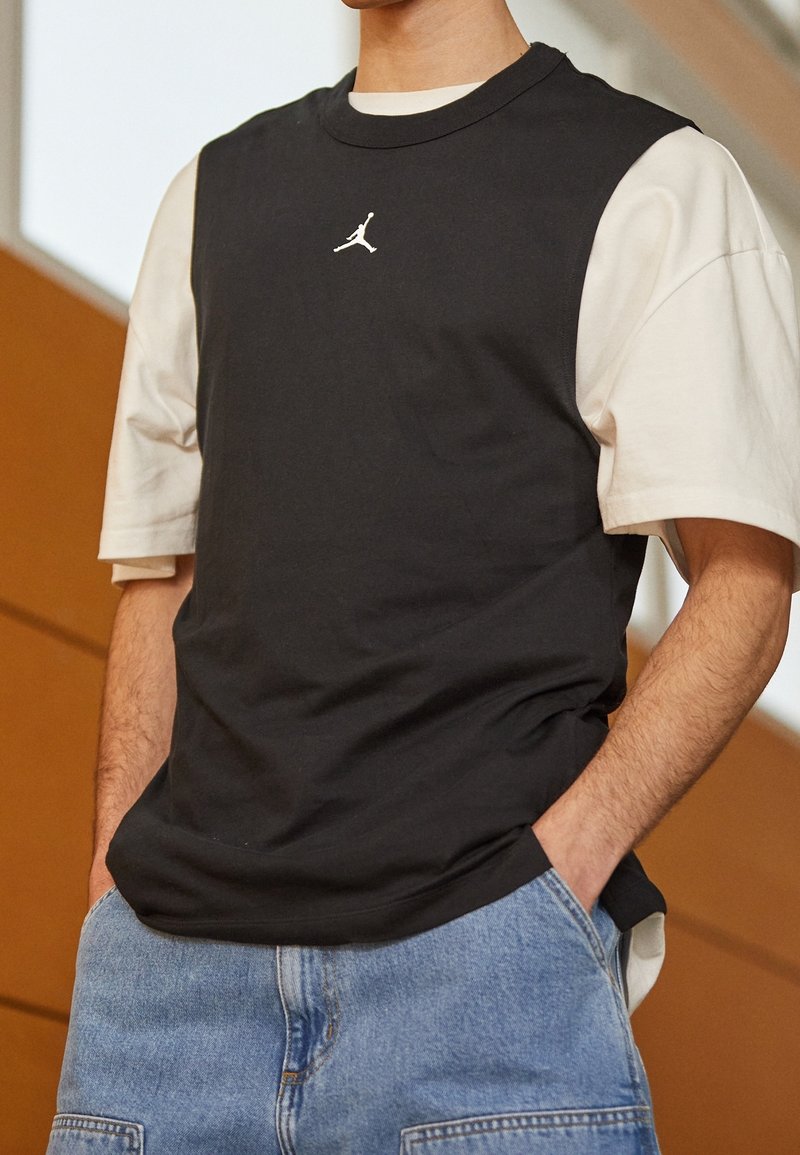 Svart ärmlös topp över en vit t-shirt. Har en liten, vit Jumpman-logotyp på bröstet. Tyget ser mjukt ut, avslappnad passform.