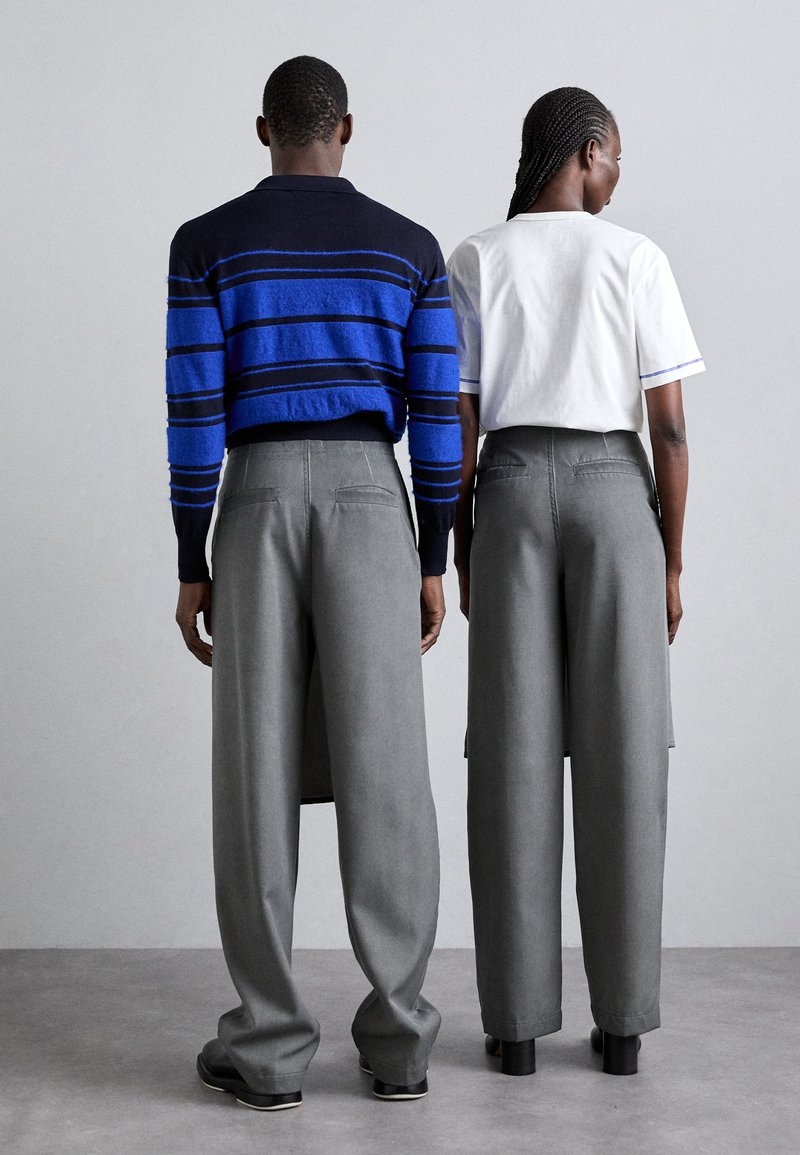 Due persone stanno schiena contro schiena. Uno indossa un maglioncino blu navy con righe blu, l'altro una maglietta bianca. Entrambi hanno pantaloni larghi grigi.